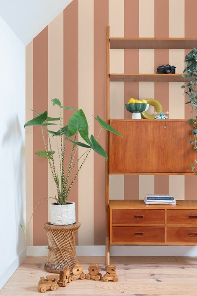 Heller Wohnraum mit breit gestreifter Tapete in Terrakotta und Beige, einem Holzregal im Retro-Stil, einer großen Zimmerpflanze und Holzspielzeug.
