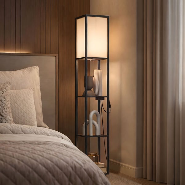Stehleuchte mit Regalböden aus Holz im Schlafzimmer neben einem Bett, rechteckiger Stoffschirm mit warmem Licht.