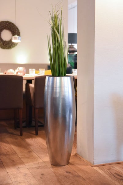 Hohe, silberfarbene Bodenvase aus Metall mit grünem Gras in einem hellen, modern eingerichteten Esszimmer mit Holzboden und Polsterstühlen.