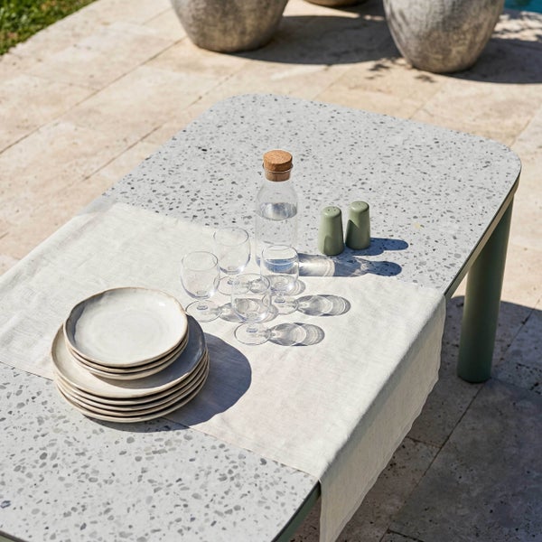 Moderner Gartentisch mit Terrazzo-Optik, gedeckt mit Keramiktellern, Gläsern und einer Karaffe auf einer hellen Steinterrasse.