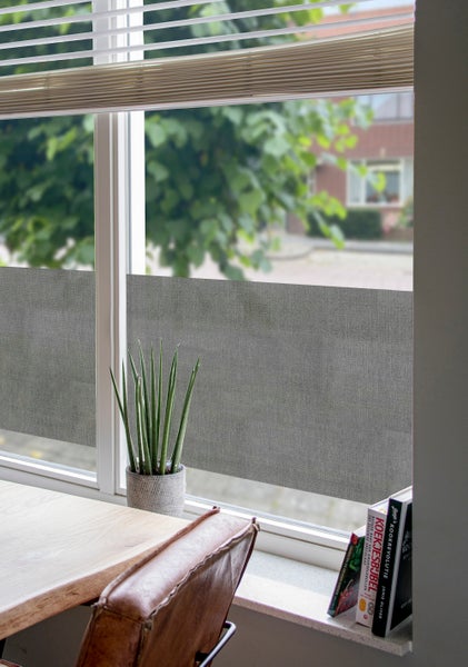 Fenster mit einer Sichtschutzfolie in grauer Leinenoptik im unteren Bereich, davor eine Zimmerpflanze und ein Holztisch.
