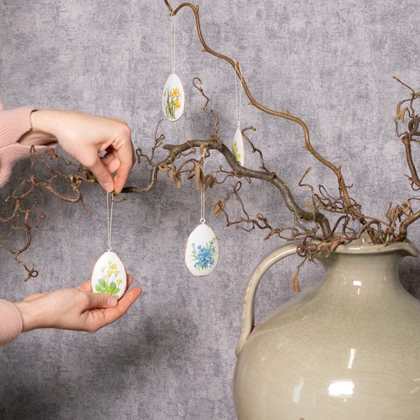 Osteranhänger in Eiform mit floralen Motiven hängend an einem Deko Zweig in einer Keramikvase mit Händen als Dekoration