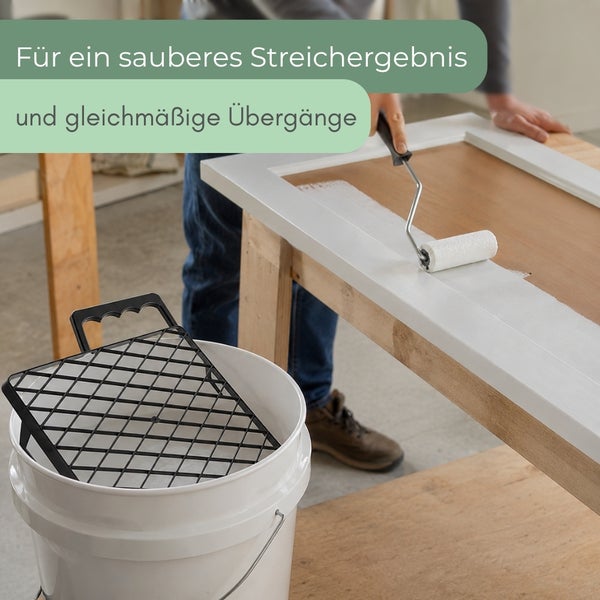 Eine Person streicht einen Holzrahmen mit einer kleinen Farbwalze, im Vordergrund steht ein Eimer mit einem Abstreifgitter.