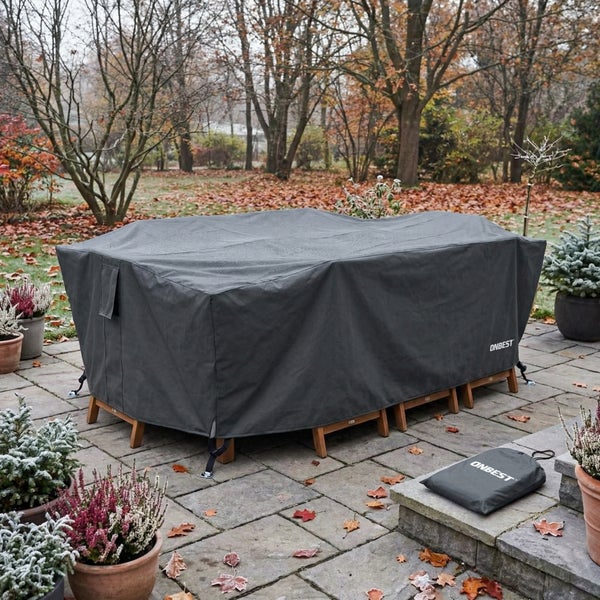 Eine graue Schutzhülle von ONBEST bedeckt Gartenmöbel auf einer gepflasterten Terrasse im winterlichen Garten mit Topfpflanzen und Bäumen.