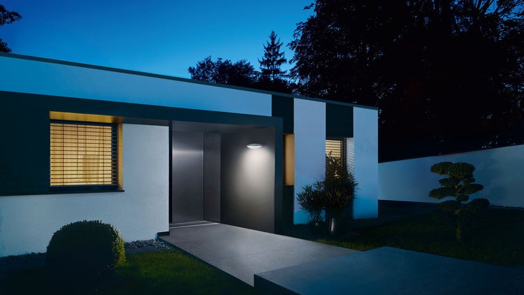 Moderner Hauseingang bei Nacht mit beleuchteter LED-Außenwandleuchte neben der Haustür und einem hellen Fenster mit Jalousien.