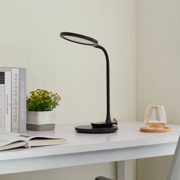 Moderne schwarze LED-Schreibtischleuchte mit rundem Kopf auf einem hellen, organisierten Schreibtisch neben Büchern und einer Pflanze.