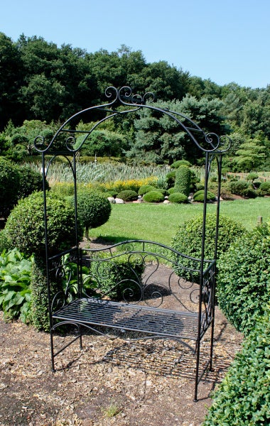 Rosenbogen mit integrierter Gartenbank aus Metall mit verschnörkelten Ornamenten in einer grünen Gartenlandschaft.
