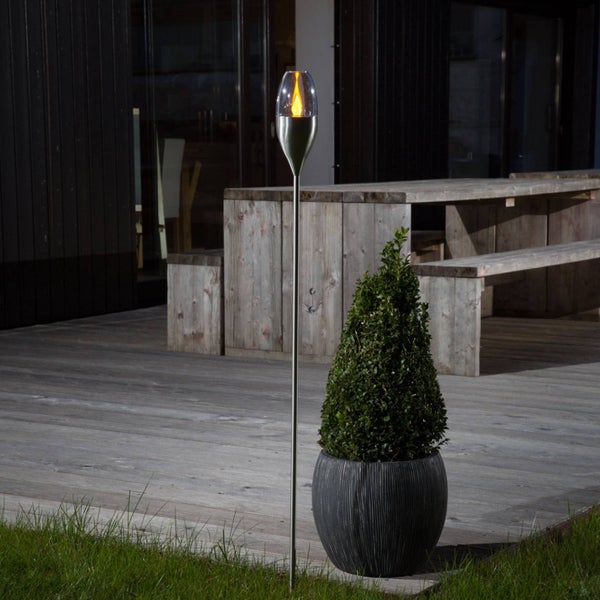 Edelstahl-Gartenfackel mit brennender Flamme in einem Glasaufsatz auf einer Holzterrasse neben einem Pflanzgefäß.