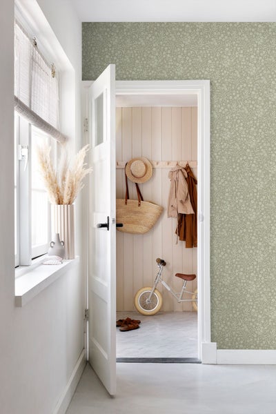 Helle Flurgestaltung mit grüner Vliestapete mit floralem Muster, weißer Tür und Dekoration aus Naturmaterialien wie Holz und Stroh.