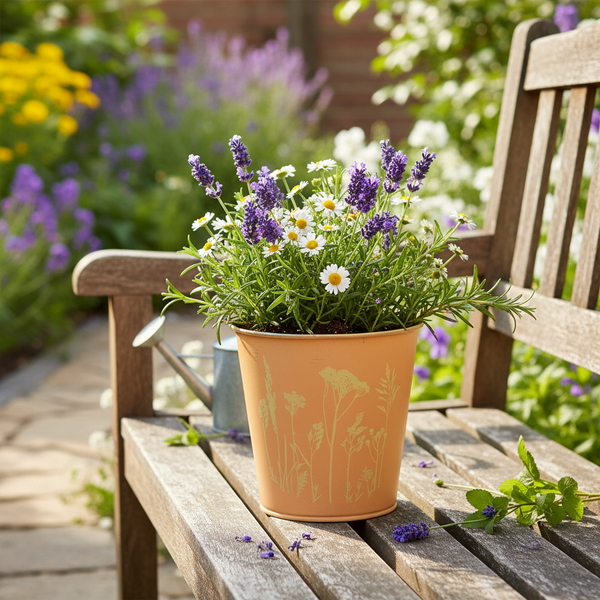 Metall-Pflanztopf mit floralem Muster, bepflanzt mit Lavendel und Gänseblümchen, auf einer hölzernen Gartenbank in einem hellen Garten.