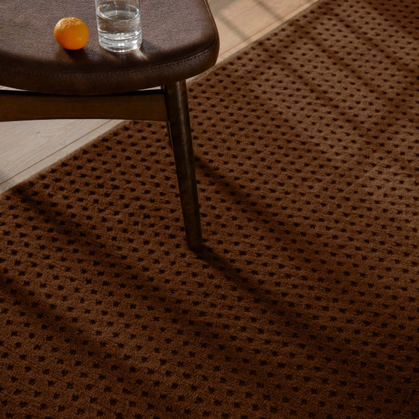 Brauner strukturierter Teppich mit Punktmuster in einem Raum mit Holzboden, einem Stuhlbein, Glas Wasser und einer Orange.