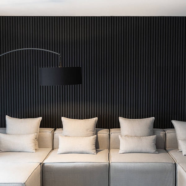 Moderne Wohnzimmerwand mit schwarzen Akustikpaneelen aus Holzlamellen hinter einem hellen Sofasystem mit Kissen und einer bogenförmigen Stehleuchte.