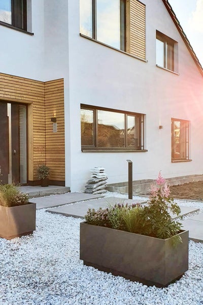 Moderner Vorgarten mit dunklen Pflanzkübeln, weißem Kies und Betonplatten vor einem hellen Haus mit Holzfassadenelementen.