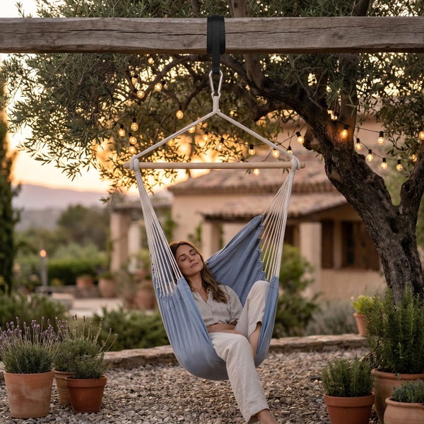 Eine Frau entspannt in einem blauen Hängesessel aus Stoff mit Holzstab, der an einem Baum in einem mediterranen Garten befestigt ist.