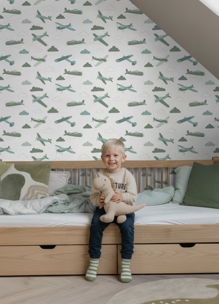 Vliestapete für Kinderzimmer mit Flugzeug- und Wolkenmotiv an einer Dachschräge, Kind sitzt auf hellem Holzbett mit Dinosaurier-Stofftier.