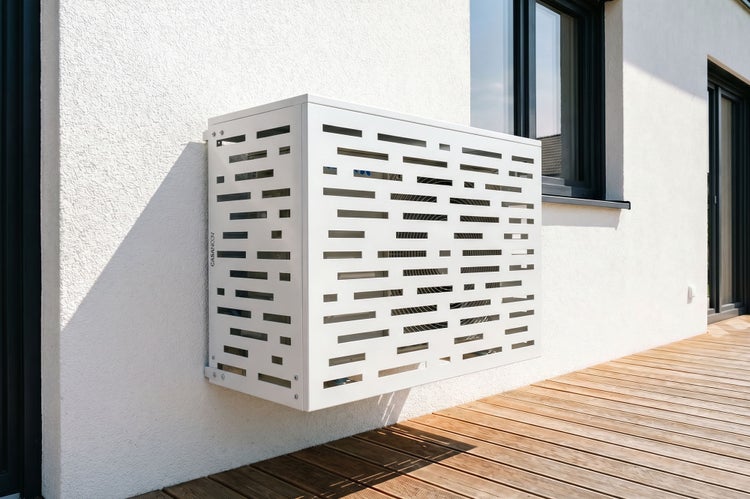 Weiße wandmontierte Klimaanlagen-Abdeckung mit Schlitzmuster an einer Hausfassade über einer Holzterrasse.