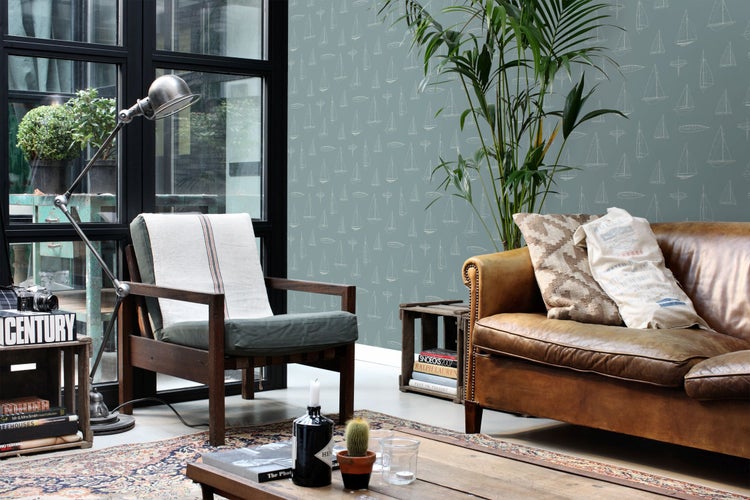 Vliestapete mit Segelboot-Muster in einem hellen Wohnzimmer im Vintage-Stil mit Ledersofa, Holzmöbeln und Zimmerpflanzen.