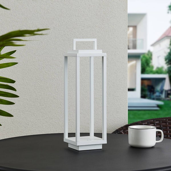 Weiße, minimalistische LED-Außenleuchte in Laternenform auf einem Tisch in heller Gartenumgebung neben einer Tasse.