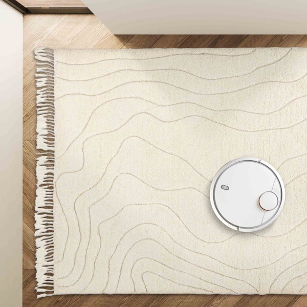 Hellbeiger Teppich mit Wellenstruktur und Fransen auf Holzfußboden, ein Saugroboter fährt darüber.