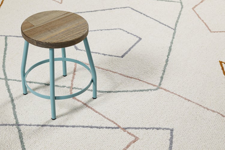 Hocker mit runder Holz-Sitzfläche und blauem Metallgestell auf einem hellen Teppich mit dezentem geometrischem Linienmuster.