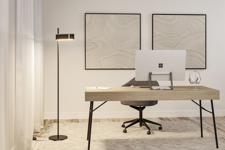 Modernes Heimbüro mit einer minimalistischen Stehleuchte, einem Holzschreibtisch, einem grauen Bürostuhl und Wandbildern in einem hellen Raum.