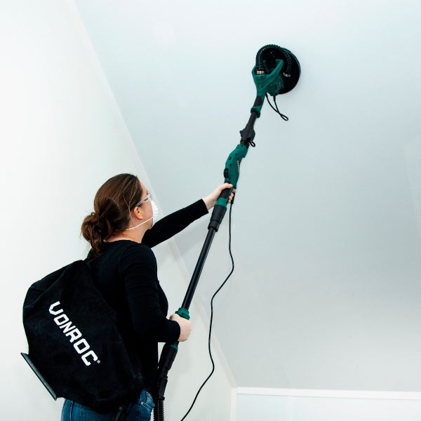 Person schleift eine Decke mit einem Teleskop-Wandschleifer von Vonroc mit Staubbeutel und persönlicher Schutzausrüstung.