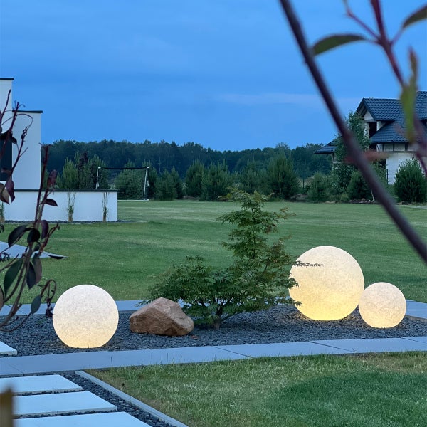 Drei leuchtende Kugelleuchten in verschiedenen Größen in einem Kiesbett mit Pflanze und Dekostein in einer modernen Gartenlandschaft.