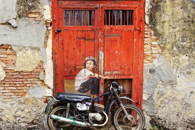 Ein altes Motorrad vor einer verwitterten roten Holztür mit einem Wandgemälde eines Kindes in einer bröckelnden Ziegelmauer