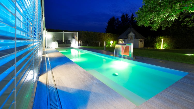 Beleuchteter Außenpool bei Nacht mit Wasserfall, Steinterrasse, Gartenhaus und stimmungsvoller Gartenbeleuchtung.