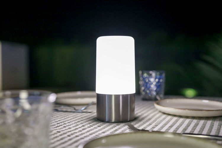 LED Tischleuchte für den Außenbereich mit zylindrischem Leuchtkörper auf einem Metallsockel auf einem gedeckten Tisch im Freien bei Nacht