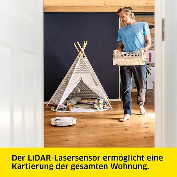 Ein Mann trägt eine Kiste in einem Raum mit einem Spielzelt und einem Saugroboter