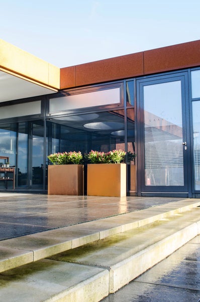 Moderne Terrasse mit zwei großen rechteckigen Pflanzgefäßen in Rostoptik, bepflanzt mit rosa Blumen, vor einer Glasfassade.