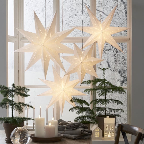 Dekoratives Arrangement mit Papiersternen, Kerzen, Weihnachtsbäumen und Lichterhäusern vor einem Fenster