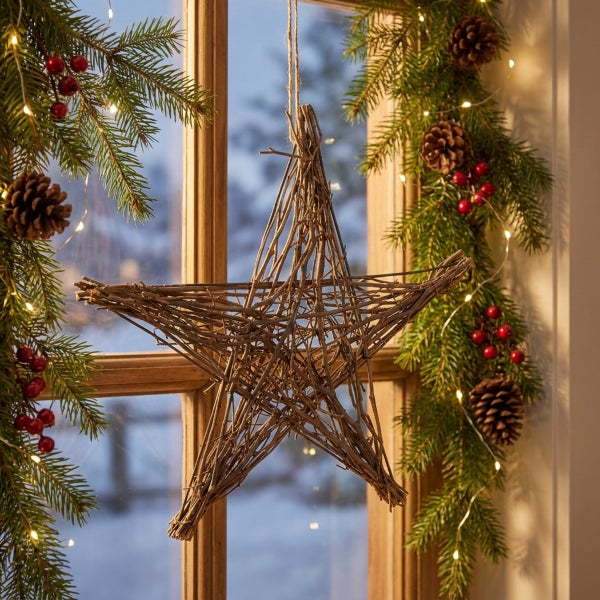 Ein dekorativer Stern aus natürlichen Zweigen hängt vor einem Fenster, dekoriert mit Tannengrün, Kiefernzapfen und einer kleinen Lichterkette.