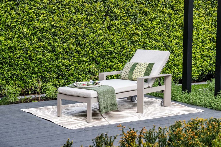 Helle Szene mit einer modernen Gartenliege aus Metall, hellen Polstern und Rollen, platziert auf einem Outdoor-Teppich vor einer grünen Hecke.