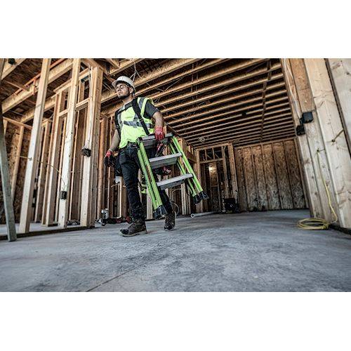 Handwerker mit Sicherheitskleidung und Schutzhelm trägt eine klappbare Arbeitsleiter durch einen Rohbau.