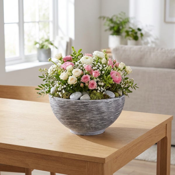 Blumengesteck in einer runden, strukturierten Schale auf einem Holztisch, arrangiert mit Rosen, Schleierkraut und Zierkieselsteinen.