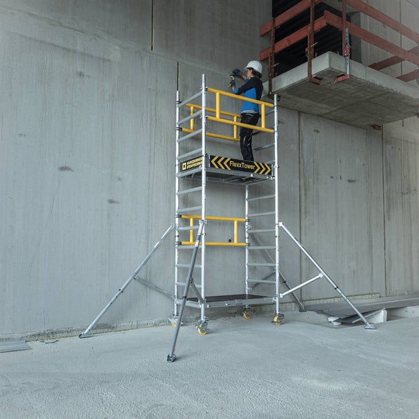 Handwerker arbeitet auf einem mobilen FlexxTower Aluminiumgerüst von Günzburger Steigtechnik vor einer Betonwand mit Sicherheitsgeländer.