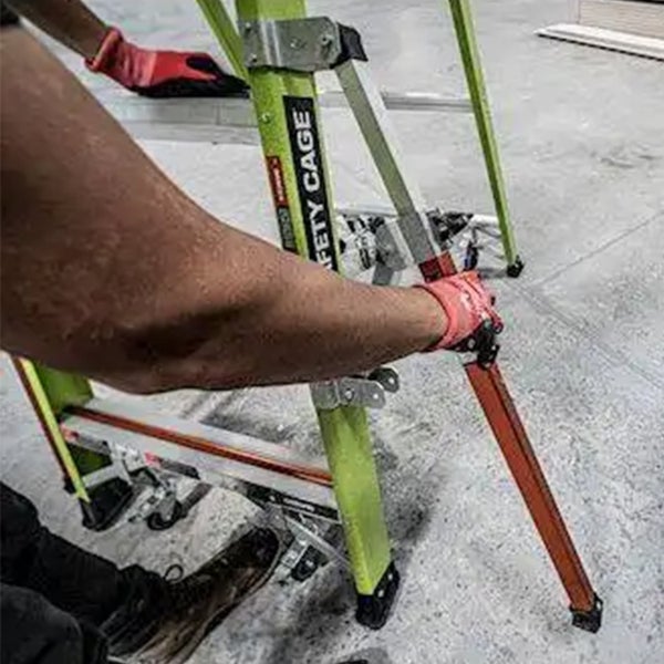 Person beim Aufbau einer Stehleiter mit Sicherheitskäfig, metallische Konstruktion mit stabilen Standfüßen und Gelenkmechanismus.