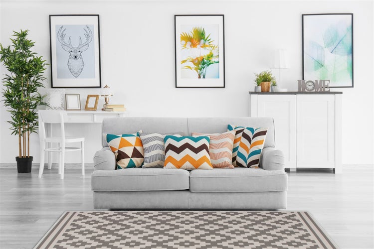 Helles Wohnzimmer mit Sofa, dekorativen Kissen, geometrischem Teppich, Zimmerpflanze und Wandbildern in einem modernen Einrichtungsstil.
