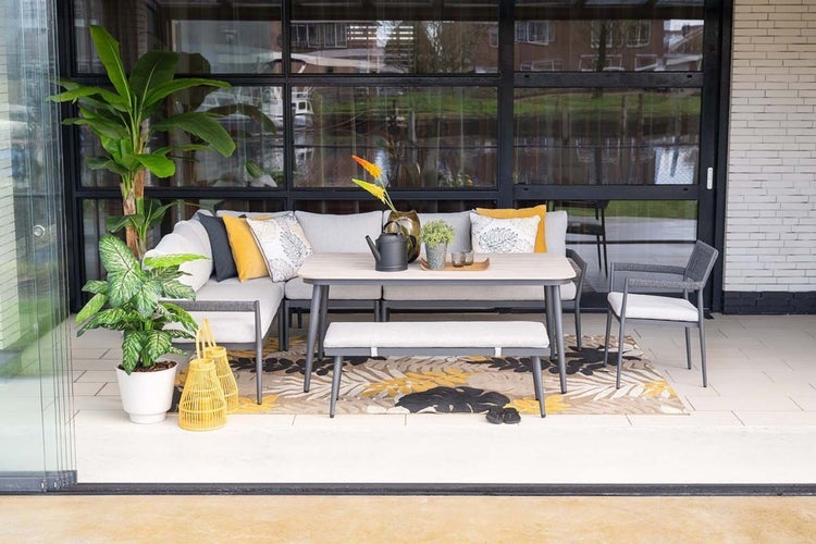 Modernes Gartenmöbel-Set auf einer hellen Terrasse mit Ecksofa, Esstisch, Sitzbank und Stuhl. Helle Polster kombiniert mit gelben Akzenten.