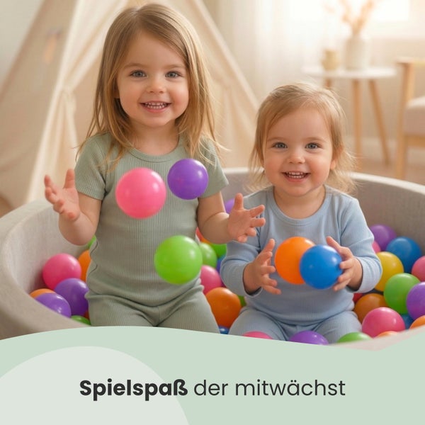 Zwei lachende Kinder spielen in einem grauen Bällebad mit vielen bunten Plastikbällen in einem hellen, freundlichen Kinderzimmer.