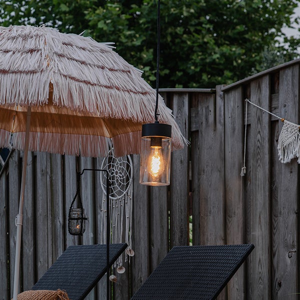 Außenhängeleuchte mit Filament-LED vor einem Fransen-Sonnenschirm und Gartenliegen an einem Holzzaun.