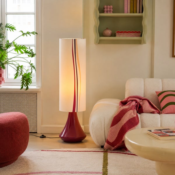 Moderne Stehleuchte mit zylindrischem Lampenschirm und geschwungenem Fuß in einem gemütlich eingerichteten Wohnzimmer mit Sofa und Pflanzen.