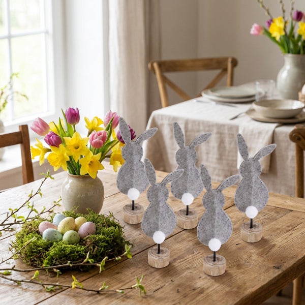 Set aus fünf Osterhasen-Dekofiguren aus Filz auf Holzständern, arrangiert auf einem Holztisch mit Frühlingsblumen und einem Nest mit Ostereiern.