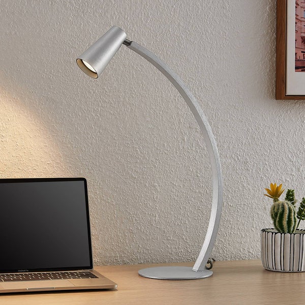 Moderne silberne Schreibtischlampe mit gebogenem Arm auf einem Holztisch neben einem Laptop und Kaktus in einem hellen Arbeitsbereich.