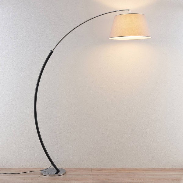 Moderne Bogenstehlampe mit gebogenem Metallgestell und beigem Textilschirm auf hellem Holzboden vor einer strukturierten Wand.