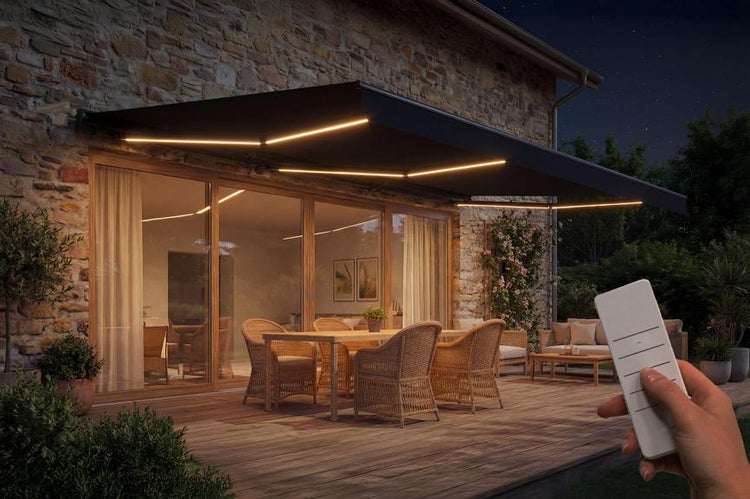 Gelenkarmmarkise mit integrierter LED-Beleuchtung an einer Natursteinwand über einer Terrasse mit Sitzgruppe bei Nacht und Fernbedienung.