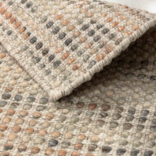 Detailansicht eines gewebten Wollteppichs mit Noppenstruktur in Beige, Grau und Braun, eine Ecke ist umgeschlagen.
