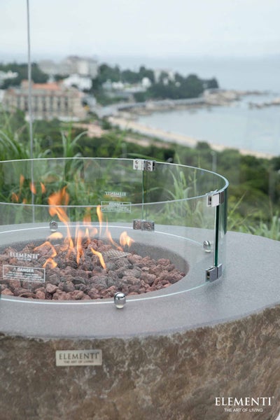 Runde Gaskaminstelle in Natursteinoptik mit Glaswindschutz, Lavasteinen und brennenden Flammen im Außenbereich.